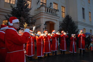 Das Foto zeigt 11 Bläser, die im Weihnachtsmann-Kostüm vor dem erleuchteten Palais Zabeltitz musizieren. Im Hintergrund sind Weihnachtsbäume zu sehen. Die Szenerie ist ansonsten dunkel.