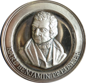 Das Foto eine silberfarbene Münze. Es handelt sich um den Avers, die Vorderseite, der Preuskermedaille. Man sieht das Konterfei von Karl Benjamin Preusker. Sein Name ist auf der Münze zu lesen.
