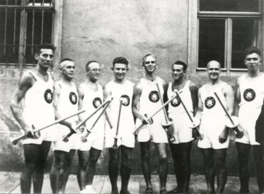 Auf einem schwarz-weiß Foto sieht man acht Männer vor einer Hausfassade, die Fackeln in den Händen tragen. Sie haben Sportkleidung an und lächeln.