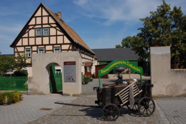 Man sieht ein ockerfarbenes Fachwerkhaus als Bestandteil eines Dreiseithofes. Im Bildvordergrund steht ein dunkelbrauner Handwagen auf dem ein grünes Schild mit der gelben Aufschrift "Bauernmuseum Zabeltitz" angebracht ist.