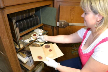 Das Foto zeigt Archivarin Anke Brekow vor einem historischen Holzmöbel. Sie trägt weiße Handschuhe und greift nach Archivalien.