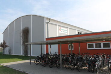 Das Foto zeigt die Sporthalle "Am Schacht". Man sieht ein großes, graues Gebäude mit halbrundem Dach und daneben ein kleineres rotes Gebäude. Davor stehen viele Fahrräder.