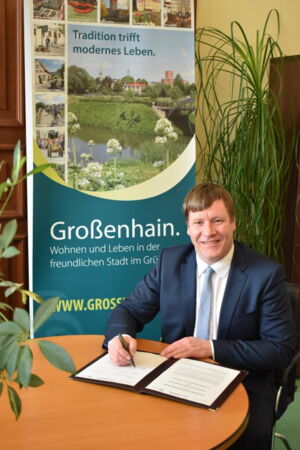 Das Foto zeigt OB Dr. Sven Mißbach sitzend an einem runden Tisch. Vor ihm liegt eine Unterschriftsmappe, darin ein Dokument zum Unterzeichnen. Er hält einen Füller in der rechten Hand und lächelt in die Kamera.
