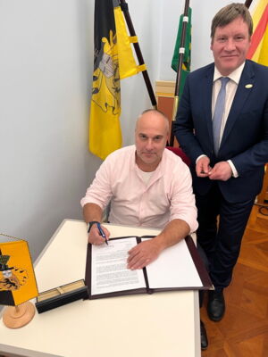 Das Foto zeigt Stadtrat Mario Gieb, sitzend, und OB Sven Mißbach, dahinter stehend. Sie lächeln in die Kamera. Vor ihnen liegt auf einem Tisch ein Schriftstück zum Unterzeichnen.