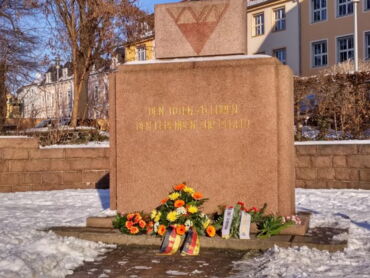 Das Foto zeigt das VVN-Denkmal an der Mozartallee, im Hintergrund ist die Berufsschule zu sehen. Vor dem Denkmal liegen Blumengebinde. Das Denkmal ist von Schnee umgeben.