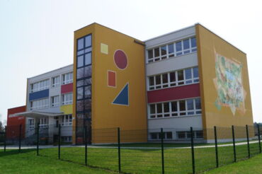 Das Foto zeigt das Schulgebäude der Grundschule Zabeltitz. Besonders die farbigen Elemente an der gelb-weißen Fassade fallen ins Auge.