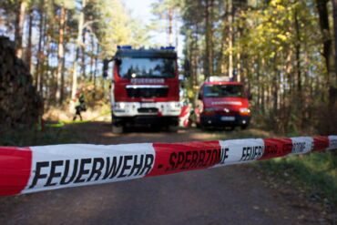 Das Foto zeigt einen von der feuerwehr gesperrten Waldbereich. Im Hintergrund sind man zwei Feuerwehrfahrzeuge.