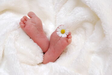 Das Foto zeigt zwei Babyfüße, die aus einer weißen Felldecke hervorschauen. Zwischen den zehen eines Fußes steckt ein Gänseblümchen.