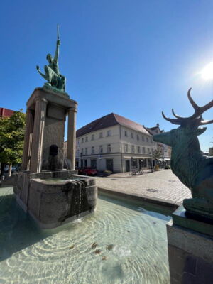 Das Foto zeigt den Dianabrunnen am Hauptmarkt. Auf der rechten Bildseite sieht man einen Hirsch. Auf Säulen in der Mitte thront die Diana mit dem Bogen. Im Brunnen befindet sich Wasser. Es scheint die Sonne.