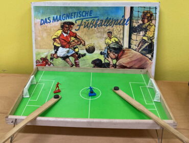 Das Foto zeigt ein kleines Fußballspiel auf einem Holzbrett. Eine rote und eine blaue Figur stehen vor zwei Toren.