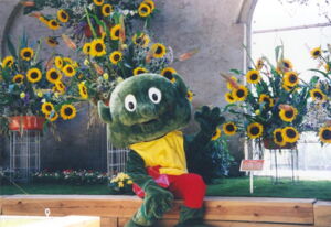 Das Foto zeigt den NIX, das Maskottchen der Landesgartenschau 2002, in Großenhain. Er sitzt vor Blumengestecken mit Sonneblumen und winkt dem Betrachter zu.