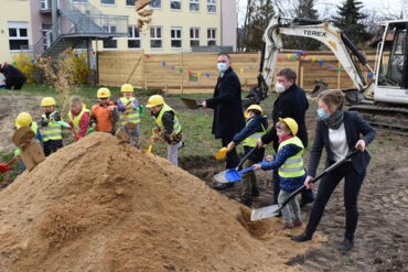 Das Foto zeigt Landrat Ralf Hänsel, Oberbürgermeister Sven Mißbach, Kitaleiterin Kathleen Michel und die Kita-Vorschulkinder wie sie Sand wegschaufeln.