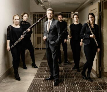Das Foto zeigt den Schauspieler Roman Knižka inmitten von Musikern des Bläserquintett OPUS 45. Sie sind alle festlich schwarz gekleidet und stehen in einem Gefängnis.