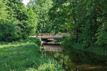 Das Foto zeigt die neugebaute Brücke. Im Vordergrund sieht man eine grüne Auenlandschaft.